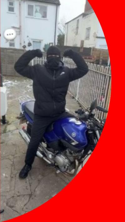 An image shows someone dressed all in black, wearing a balaclava, posing with their arms in the air as they sit on a motorbike