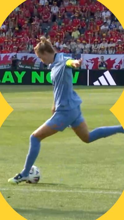 Vivianne Miedema wears blue Netherlands kit as she prepares to strike football in stadium. Behind her are crowd wearing red Wales football shirts.
