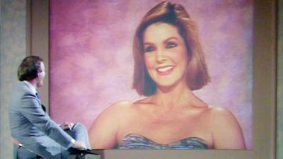 Wogan sitting in front of Priscilla Presley who is speaking from a large screen behind him.