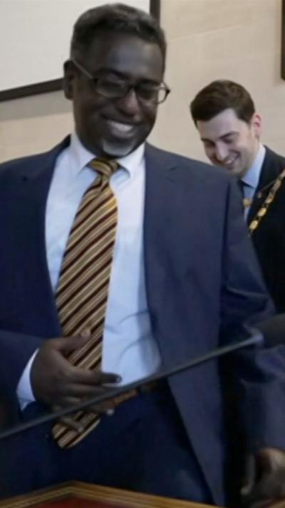 Yassin Mohamud is standing and smiling. He is wearing a blue suit, white shirt and a yellow and claret tie. He is inside a council chamber and the former Lord Mayor is in the background, he is also smiling.