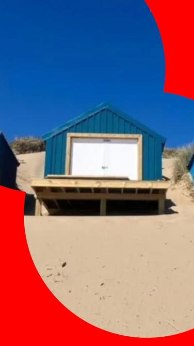 A teal-coloured beach hut with white double doors, sited at the top of a sand bank.