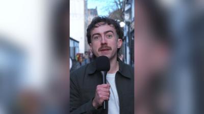 Jack Doherty standing in Glasgow's Ashton lane holding a black microphone. He has short dark hair, a moustache and is wearing a dark grey collared jacket and a white t-shirt.