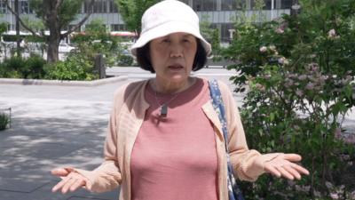 A woman in a pink shirt and orange cardigan holds her hands out in a questioning pose.