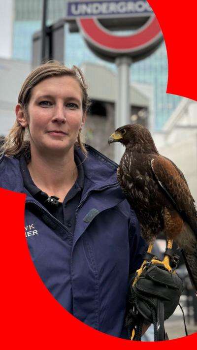 A falconer in a dark jacket, holding a hawk, stands near a London Underground sign amidst a cityscape of modern buildings