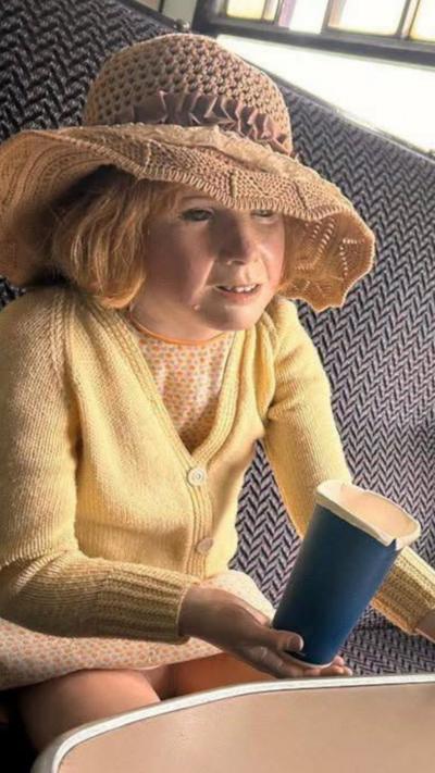Child mannequin in a yellow cardigan and straw hat sitting on a train seat holding a blue paper cup
