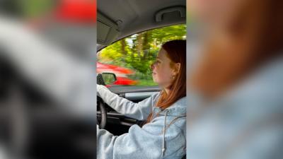 A young female driver with medium length red hair driving in a car. She is wearing a light grey hoody and there are trees visible out the driver's window and a passing red car.