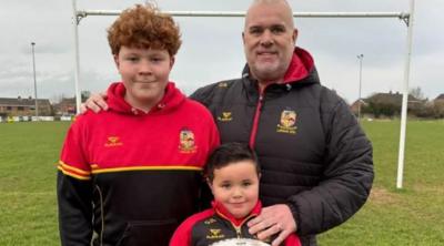 Ollie smiles holding a red and white rugby ball at Lurgan rugby pitches. His dad Colin has his hand on Ollie's shoulder and is wearing a black coat. Colin also has his arm around his son Niall who has red hair and is smiling. Niall is wearing a red and black hoody.