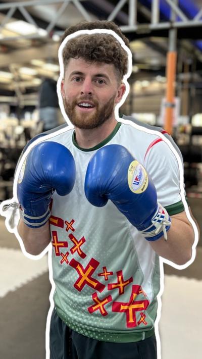 A man with brown hair and a beard looking at the camera holding up his fists in blue boxing gloves