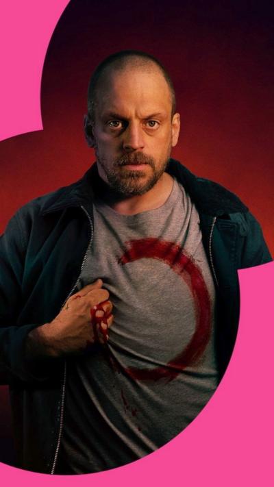 Actor Matthew Gurney wearing a T-shirt with a red unfinished circle and his bloodied hand on his chest, against red background