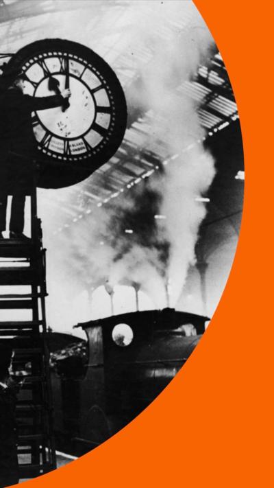 A black and white image of a man manually changing a clock. A steam train can be seen in the background.