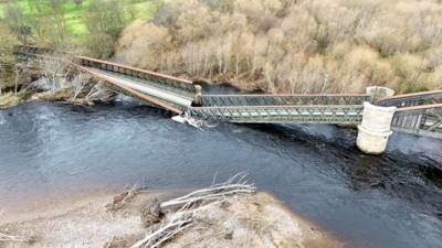 Drone image of a collapsed iron bridge, twisted and snapped in the middle, lying in a river.