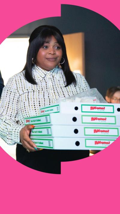 A woman in a white patterned shirt holds four pizza boxes