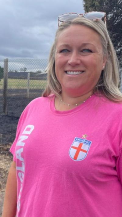 A woman is wearing a pink t-shirt and has blonde hair, she is standing in the middle of a charred field. 