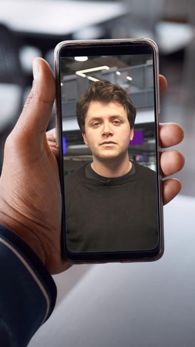A man with brunette hair is looking towards the camera and is in a phone held by a hand.