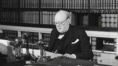A black and white photo of Winston Churchill wearing glasses, sat behind a desk with microphones
