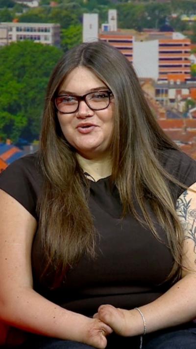 A teenager with long dark hair and glasses on a TV studio sofa