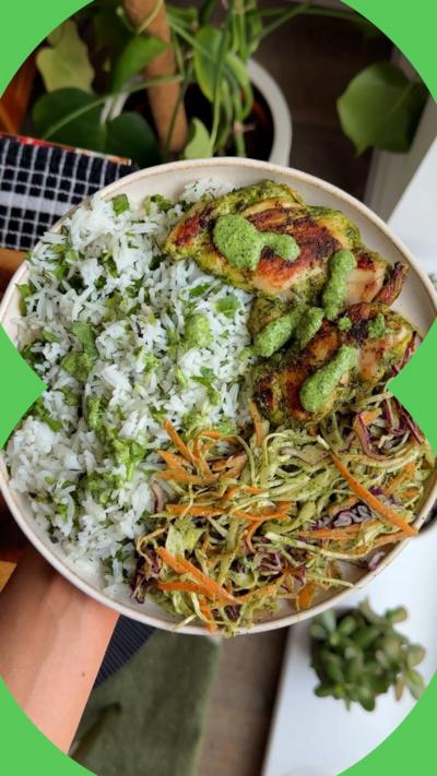 A hand holding a white plate filled with browned chicken, fresh slaw and herby rice, with plants in the background