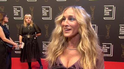 Sarah Jessica Parker, with long wavy blonde hair and a pale purple dress, smiles on the red carpet.