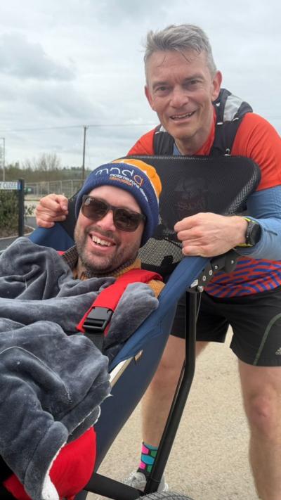 A man in running gear huddles over a man in a sports wheelchair. They are both smiling at the camera.
