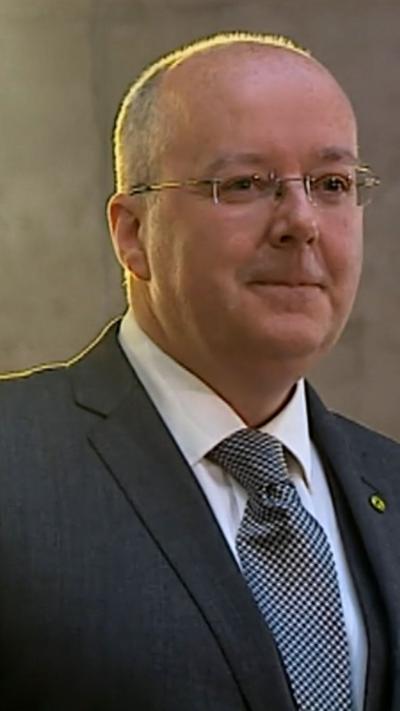 Former SNP chief executive Peter Murrell smiles as he looks off to the side. He is wearing glasses, a white shirt, dark grey blazer and a black and white tie.
