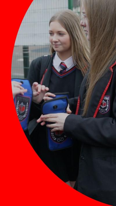 Two female pupils stand holding blue pouches containing their mobile phones.