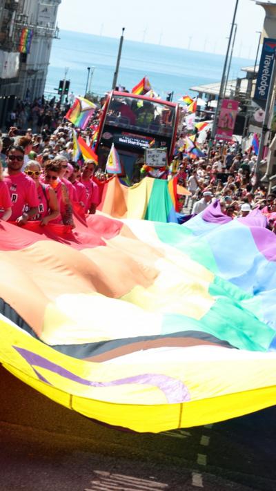 People holding a large LGBTQ+ flag in a parade.
