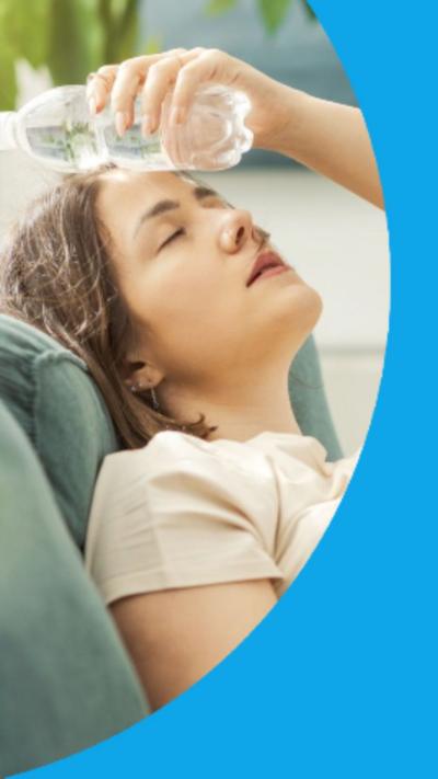 A woman reclines on a sofa next to a fan and with a bottle of water pressed to her forehead