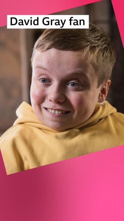 A boy smiles in front of a fireplace - he has short dark blond hair and wears a yellow hoodie