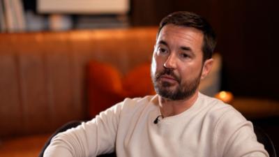 Martin Compston faces the camera, he wears a cream top, has dark hair and a beard, the room behind him is in soft focus