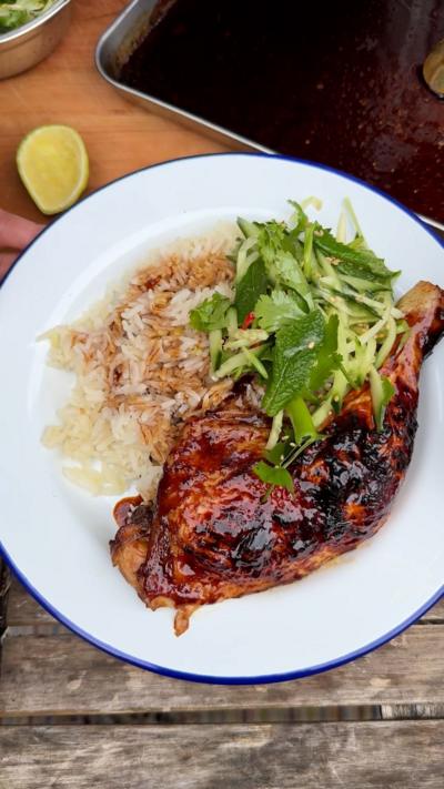 A cooked, glazed chicken leg on a white enamel plate with rice and green slaw, on a wooden table