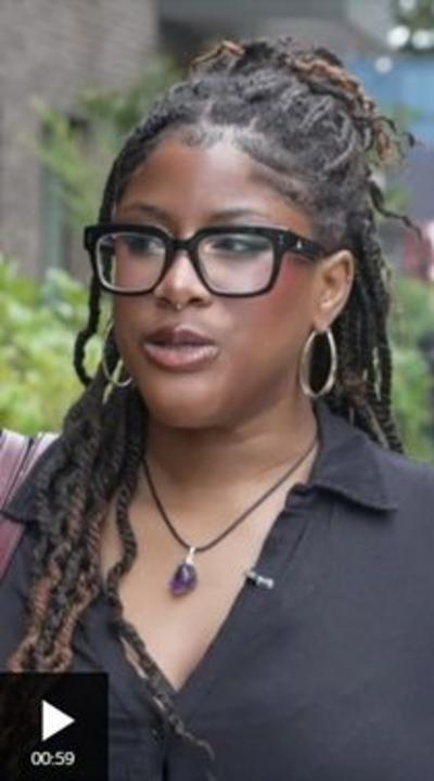 Woman with dreadlocks, black shirt, and purple necklace standing on a sidewalk near a brick building and greenery.