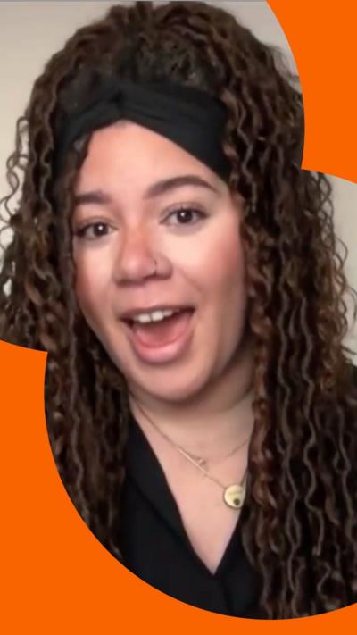 A woman with long brown curly hair in braids talks to the camera. She is wearing a black top and a black hairband.