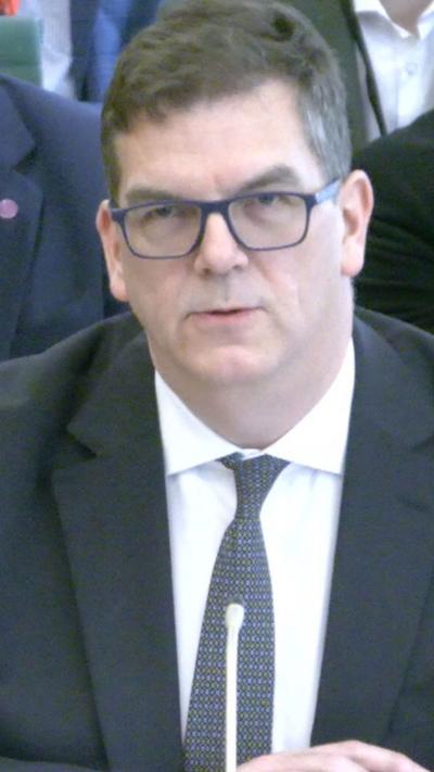Olly Robbins speaks at a microphone wearing a black suit