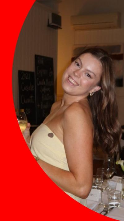 A young woman with long brown hair smiles while holding a glass of wine. She is wearing a strapless yellow dress.