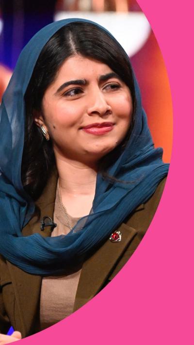 Malala Yousafzai smiles in the Graham Norton Show studio. She has long black hair and wears a blue headscarf and brown jacket