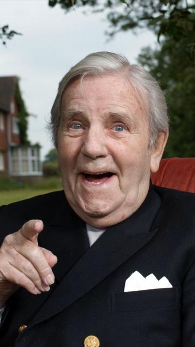 An elderly Norman Wisdom outdoors pointing to camera smiling
