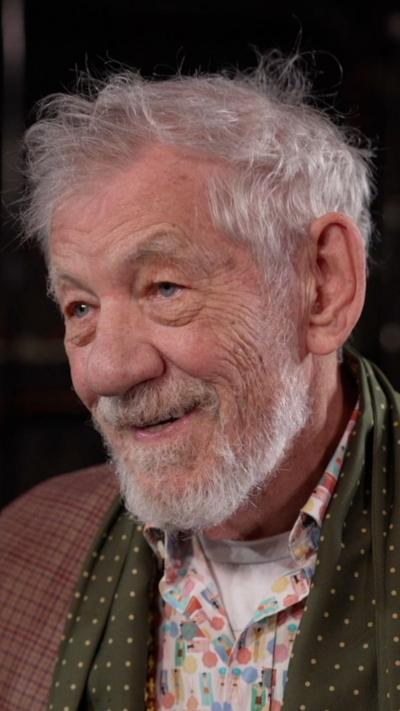 Sir Ian McKellen looking off camera. He is smiling and has grey hair and a beard. He is wearing a patterned shirt and scarf with a brown checked jacket.