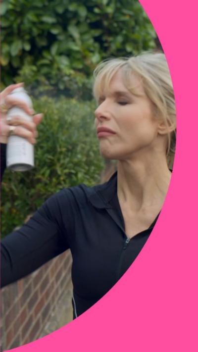 Lucy Punch as Amanda is wearing a black gym top and spraying a bottle of makeup setting spray on her face.