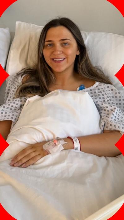 A woman with brown hair sits in a hospital bed. She wears a gown and there is a catheter in her left hand