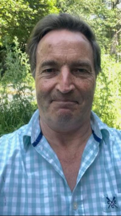David Braine in a blue and white top. He is smiling and looking directly at the camera. He has brown hair. Behind him are green trees.