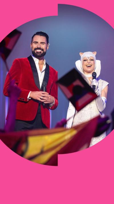 Rylan and his co-host of the Interstellar Song Contest in Doctor Who stand on stage as people wave flags in the air