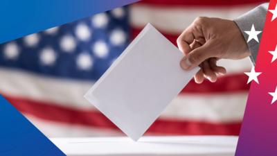 A hand puts a ballot into a ballot box with a US flag in the background