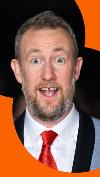 Alex Horne has brown hair and a moustache, and a greying beard. He is wearing a white shirt, red tie, and a two-tone blazer.