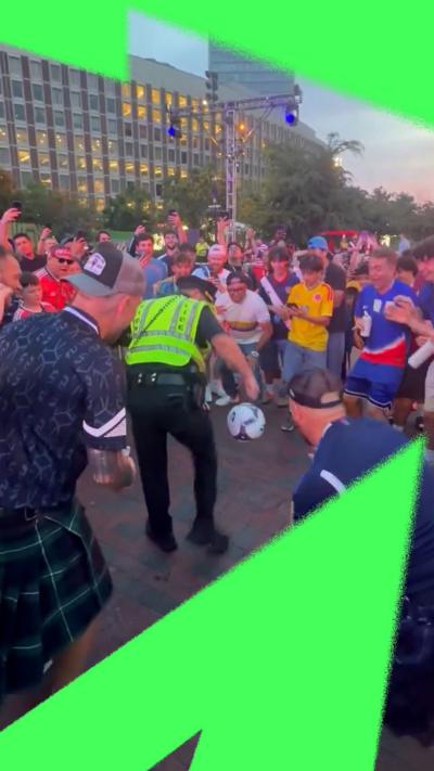 A police officer is kicking a football surrounded by Scottish football fans cheering.