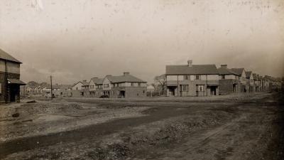 An old black and white image of a housing estate being built. Muddy roads lead towards houses with a grey sky overhead.