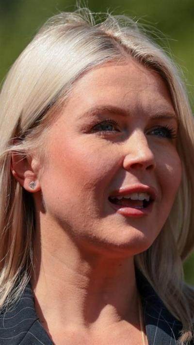 A blonde woman speaks off towards the left of the camera with greenery in the far background.