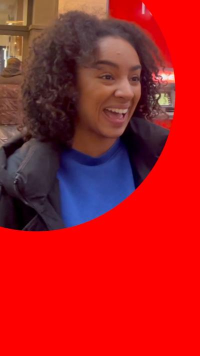 A woman with curly black hair laughs in the street while talking into a mic