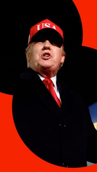 Donald Trump wearing a black jacket, red tie, and a read hat which says 'USA' on it.