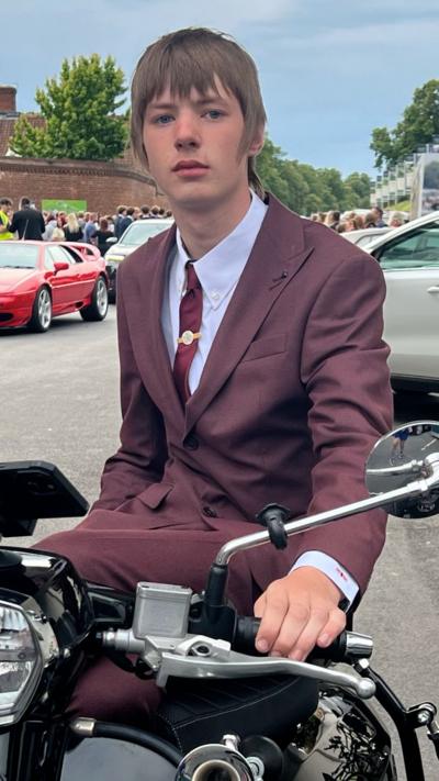 Teenager with Mod haircut wears a damson coloured suit and sits astride a motorised scooter.