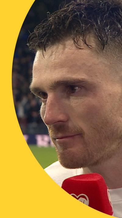 Andy Robertson has short brown hair and wears a white top. He is on a football pitch